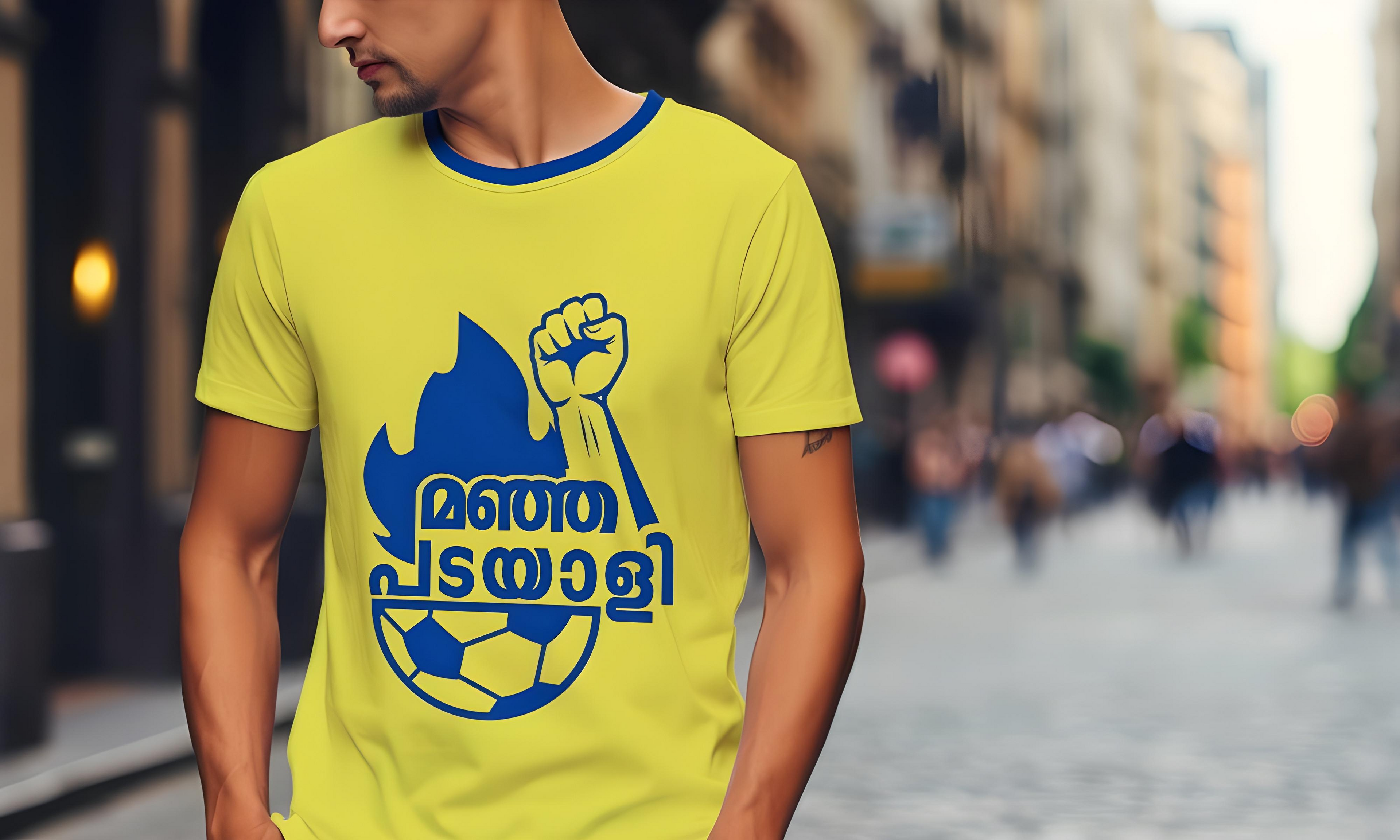 A man wearing a bright yellow T-shirt featuring bold blue artwork. The design includes a raised fist emerging from a blue flame, a stylized football, and Malayalam text that reads "മഞ്ഞ പടയാളി" (Manja Padayali), which translates to "Yellow Warrior" — a slogan associated with Kerala Blasters FC fans. The background shows a blurred city street with people walking.